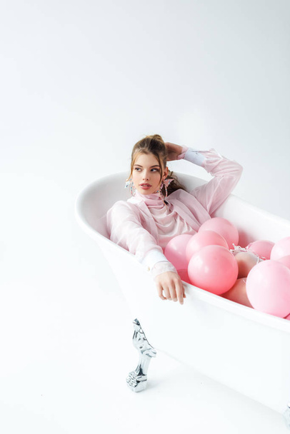 pretty young woman lying in bathtub with pink air balloons on white  - Фото, изображение