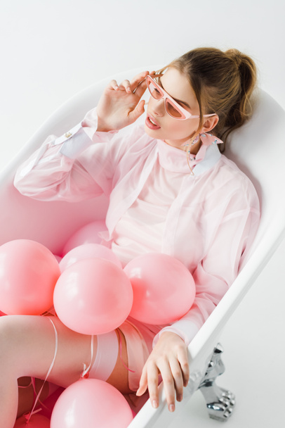 overhead view of attractive girl touching sunglasses while lying in bathtub with pink air balloons on white  - Foto, immagini