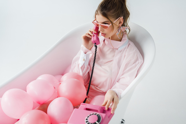 attractive girl in sunglasses talking on retro phone while lying in bathtub with pink air balloons on white  - Photo, Image