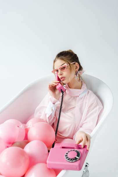 girl in sunglasses talking on retro phone while lying in bathtub with pink air balloons on white  - Photo, Image