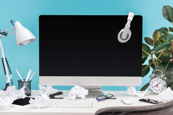 local de trabalho confuso com fones de ouvido no computador, lâmpada, papéis amassados e artigos de papelaria na mesa branca no fundo azul
 - Foto, Imagem