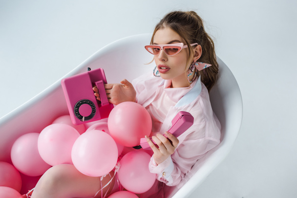 young woman in sunglasses looking at pink retro phone while lying in bathtub with air balloons on white  - Valokuva, kuva