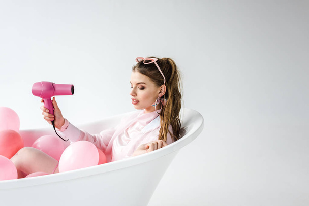 pretty girl using hair dryer while lying in bathtub with pink air balloons on white  - Foto, afbeelding