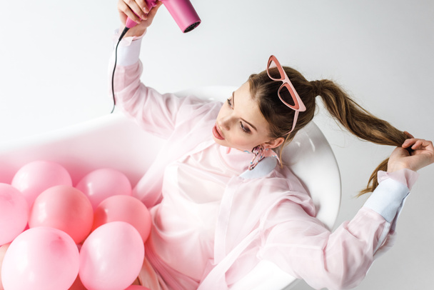 overhead view of pretty girl using hair dryer while lying in bathtub with pink air balloons on white  - Fotografie, Obrázek