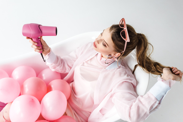 overhead view of girl using hair dryer while lying in bathtub with pink air balloons on white  - Valokuva, kuva