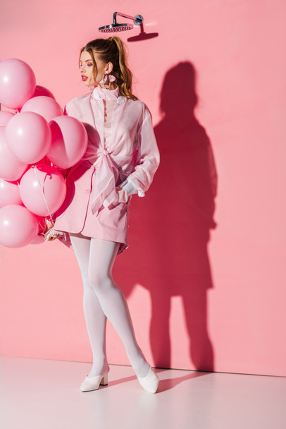 όμορφη νεαρή γυναίκα κρατώντας αέρα μπαλόνια ενώ στέκεται με το χέρι στην τσέπη για ροζ - Φωτογραφία, εικόνα