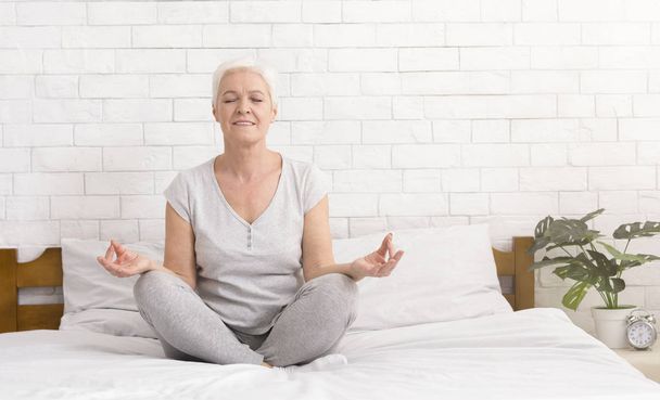 Старша жінка, що медитує в позі лотоса на ліжку
 - Фото, зображення