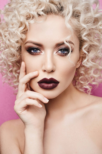 Beautiful and fashionable model girl with amazing blue eyes, with curly blonde hair and with dark red lips isolated at the pink background - Zdjęcie, obraz