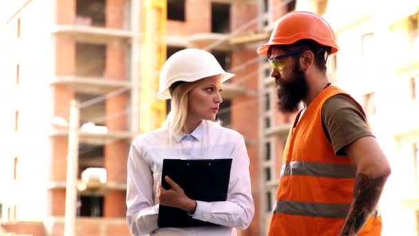 Két civil mérnökök gazdaság tervek és ellenőrzést az építkezés. Férfi és női építő védősisakok kommunikálni a háttérben az építés alatt álló épület. - Felvétel, videó
