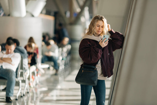Curly blonde woman with phone in hands standing in waiting room at airport. - Foto, afbeelding