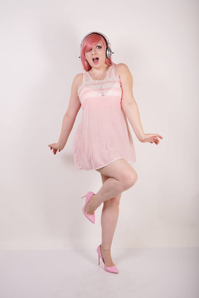 sexual plump redhaired girl wearing pink lingerie babydoll dress with headphones and having cosplay fun on white studio background - Foto, imagen