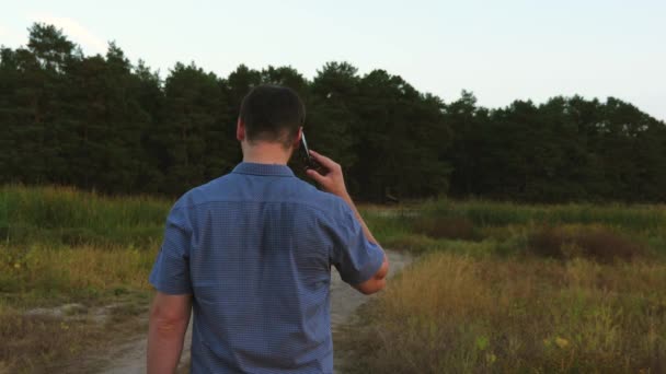 man traveling on a forest road and talking on a smartphone. tourist in the forest. man is talking on phone while walking in park in the evening. - Video, Çekim