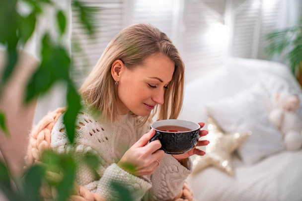 blonde fille dans un pull avec une tasse de thé assis sur le lit
 - Photo, image