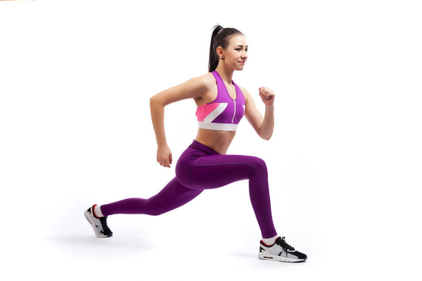 Una mujer de cabello oscuro entrenador en una camiseta corta de color rosa deportivo y leggings gimnasio hace saltos por los pies hacia adelante, las manos se extienden hacia un lado en un fondo blanco aislado en el estudio
  - Foto, Imagen