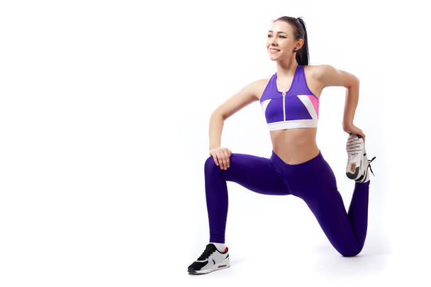 A dark-haired woman coach in a sporty purple  short top and gym leggings makes lunges  by the feet forward,  stretch legs on a  white isolated background in studio  - Photo, Image