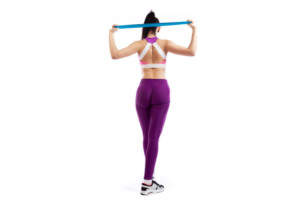 A dark-haired woman coach in a sporty purple  short top and gym leggings exercise for your back, hands behind his head breeding with sport fitness rubber bands on a  white isolated background in studio  - Фото, зображення