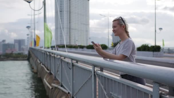 Slim woman with a pigtail blonde in a gray T-shirt standing on the bridge and using smartphone over background sea and city. Girl touching screen - Video, Çekim