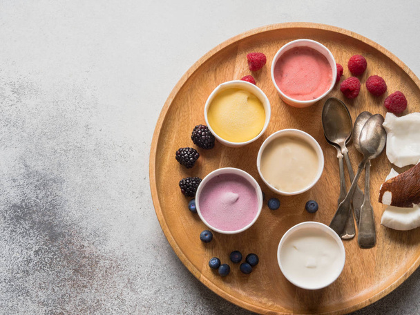 Свежие ягоды мороженое разных цветов в бумажных стаканчиках и различные ягоды на круглом деревянном подносе на сером фоне. Вид сверху. копировальное пространство
 - Фото, изображение