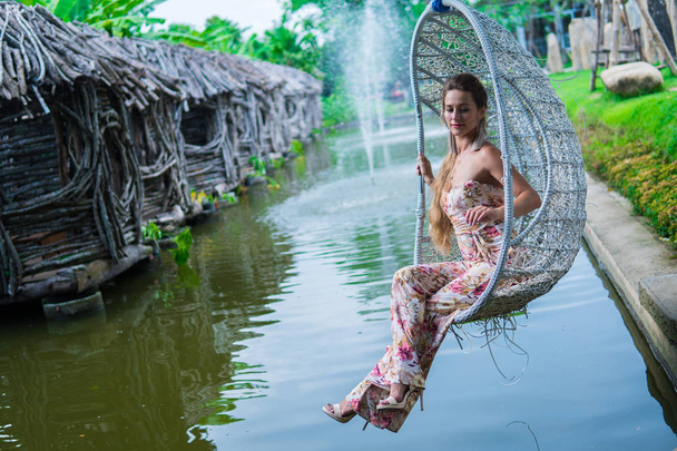 Hermosa morena en vestido balanceándose en un columpio sobre el agua en el Parque en día soleado
 - Foto, imagen