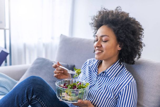 Hermosa mujer afroamericana comiendo ensalada en el sofá en la sala de estar
 - Foto, imagen