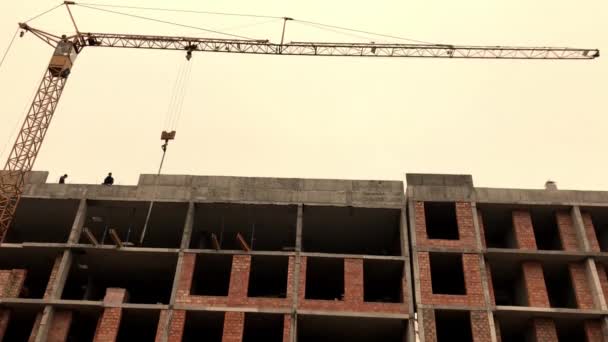 Bau eines mehrstöckigen Backsteingebäudes. Ein Kran hebt Baumaterialien. Bauarbeiter auf dem Dach des Gebäudes. - Filmmaterial, Video