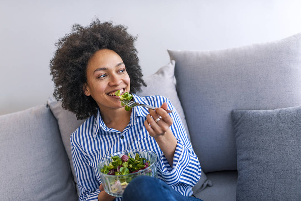 Beautiful African American woman eating salad on sofa in living room - Foto, Bild