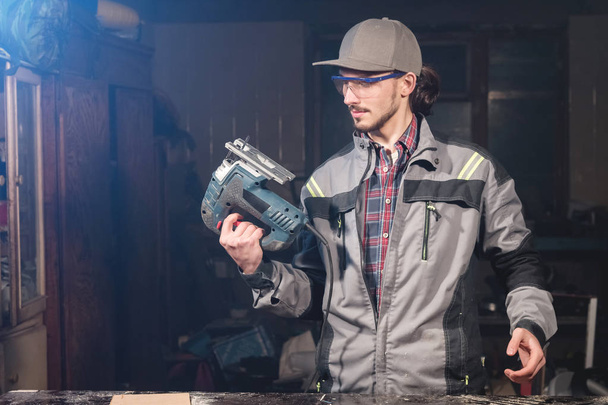 Portrait of a young carpenter joiner in overalls wearing a cap and goggles with an electric jigsaw in hand at a home workshop. Starting a business. Craftsman - Foto, afbeelding
