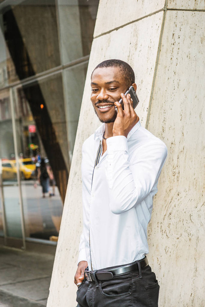 Appelle dehors. Jeune homme d'affaires afro-américain barbu, vêtu d'une chemise blanche, debout dans la rue devant un immeuble de bureaux à New York, regardant en bas, souriant, parlant sur un téléphone portable
 - Photo, image