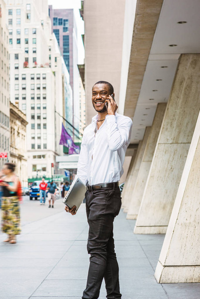 Jovem empresário afro-americano viajando, trabalhando em Nova York, vestindo camisa branca, calças pretas, carregando computador portátil, saindo do prédio de escritórios, conversando no celular, sorrindo
 - Foto, Imagem