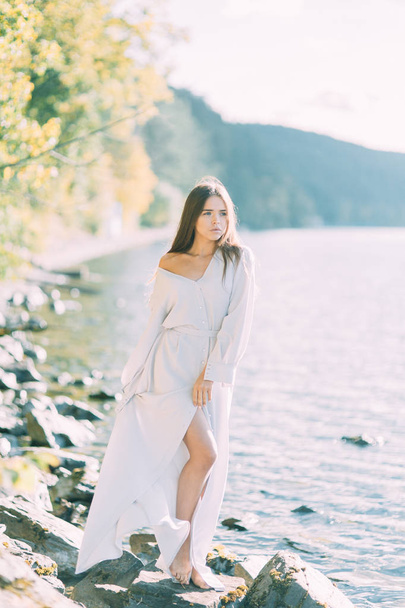 Girl in boudoir dress on the lake, standing and smiling. Unity with nature, morning bride in Russia. Beautiful eyes and model appearance - Fotó, kép
