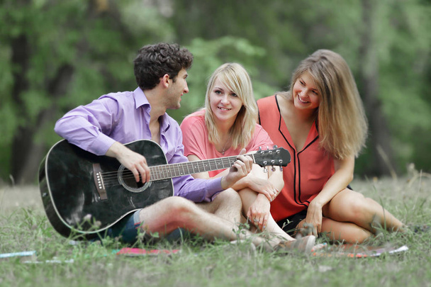 τρεις φοιτητές με μια κιθάρα που κάθεται στο γρασίδι στην πόλη πάρκο - Φωτογραφία, εικόνα