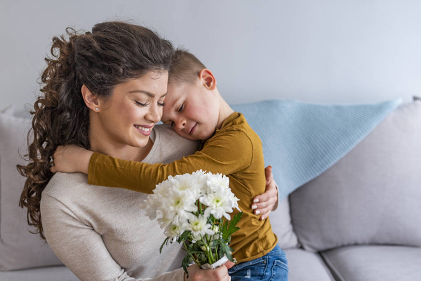 Mother and little son with bouquet of flowers hugging on sofa - Photo, Image