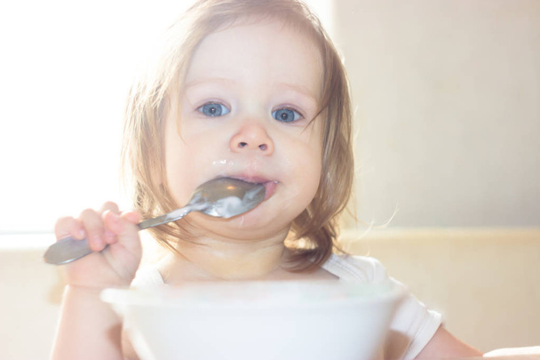 Little shaggy baby girl with pleasure alone holds a spoon and eats delicious food from a white plate. - Photo, Image