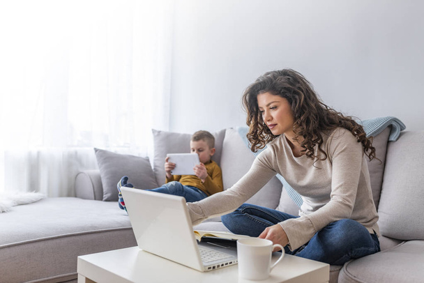 Mother working on laptop at home while son using tablet on sofa - Fotó, kép