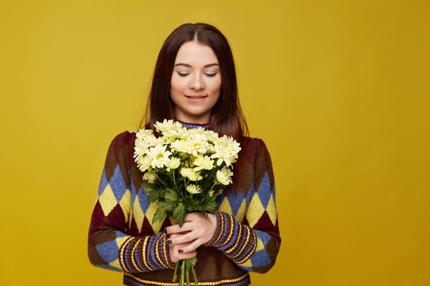 Gorgeous European woman with healthy clean skin and beautiful set of features, dressed in knitted jumper, wearing hair loose, biting lip with shy charming smile, looking down at l bouquet of flowers. - 写真・画像