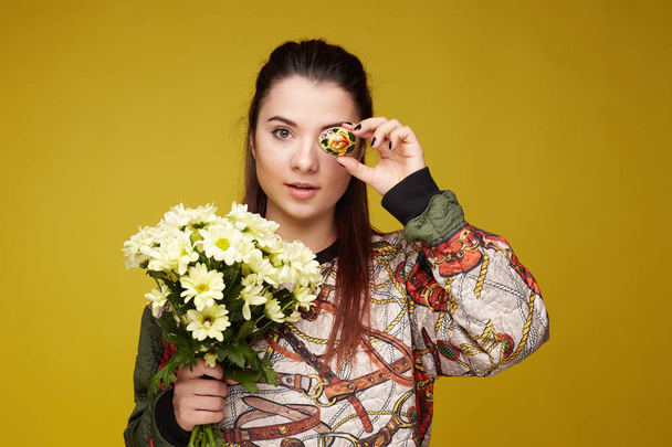 Attractive Caucasian woman covers eye with Easter egg, wears colorful outfit, prepared delicious traditional dish for holiday, models over yellow wall background with free space for promotion content. - Photo, Image