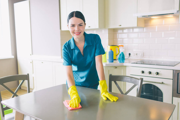 Smiling cleaner is standing and looking on camera. She cleans table. Girl wears special blue uniform. Woman works in apartment. - Foto, afbeelding