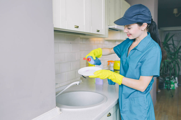 Cleaner in blue uniform and cap holds plate in hands. She pours liquid for cleaning dishes on plate. Girl in concentrated. She smiles. - Foto, immagini