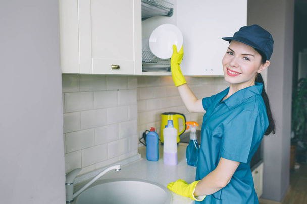 Positive and energetic girl stands at sink and look on camera. She smiles. Woman puts plate in cupboard. She looks careful. Girl is professional cleaner. - Fotoğraf, Görsel
