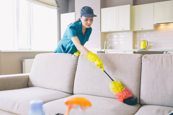 Careful and concentrated cleaner works in apartment. She uses dust brush on sofa. Girl cleans gentle. She is reaching far angles of sofa. - Photo, image