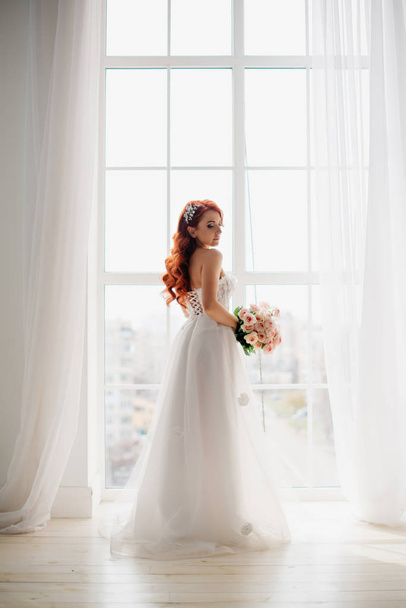 Red-haired bride in white wedding dress stands near large window - Фото, зображення