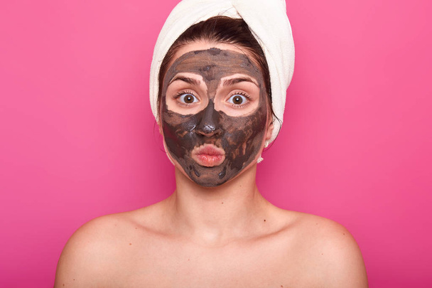 Close up shot of beautiful female, has chocolate mask on face, keeps eyes wide opened and lips rounded, poses with naked body, has white towel on head, having rest in spa salon, isolated on pink wall. - Fotoğraf, Görsel