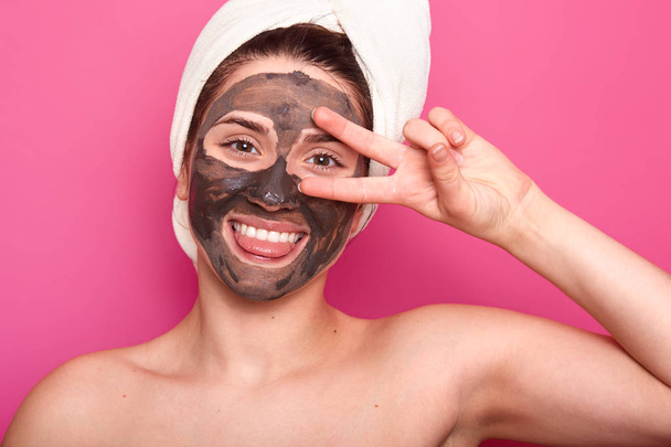 Overemotive pleased woman with toothy smile, shows her tougne, keeps two fingers near her eye, rejoices smooth healthy skin, wears white towel on head, isolated on pink background. Skin care concept - 写真・画像
