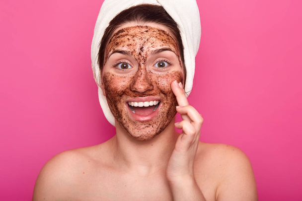 Isolated shot of surprised woman applies mask on face, has happy facial expresion, opens mouth from amezmant, having rest while doing beauty procedures, wears white towel on head. Wellness concept - Фото, изображение