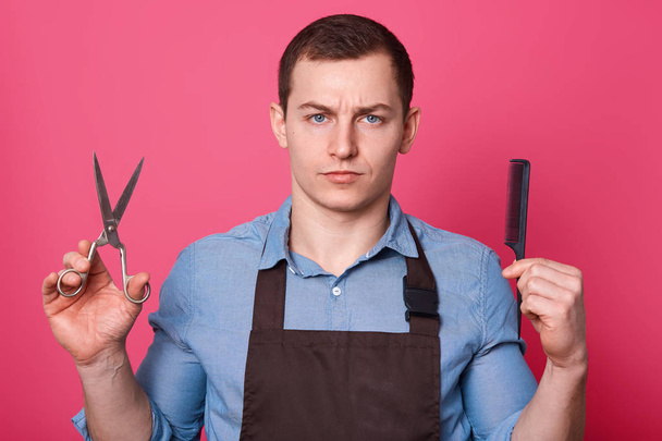 Close up portrait of handsome barber with serious facial expression, holds scissors, hair comb, ready for making hairstyle, looks at camera, dressed in shirt and apron, isolated over pink background. - Фото, зображення