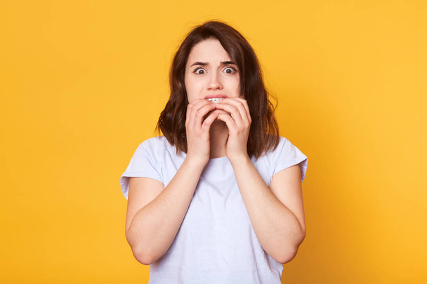 Portrait of frightened brunette girl bites fingers nails, has frightneed facial expression, afraids of something, wears in casual white t shirt, models on yellow background. Negative feelings concept - Photo, image