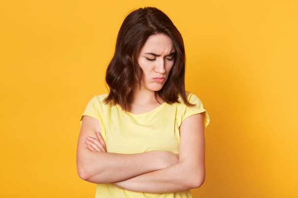 Portrait of upset unsatisfied Caucasian woman standing with arms folded and looking down isolated over yellow background, has dark wavy hair, looks offended. People and relation concept. Copy space. - Fotografie, Obrázek