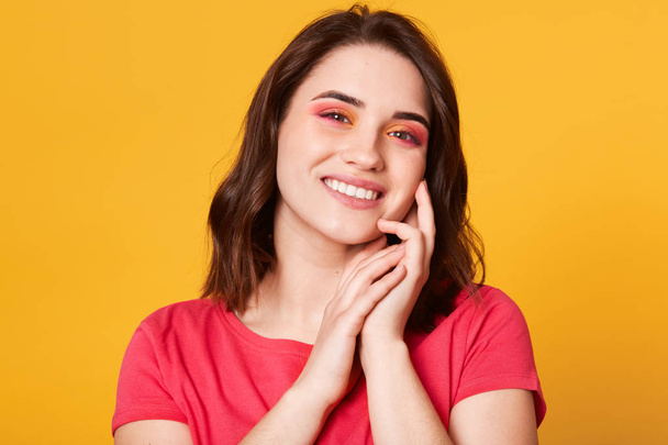 Image of cute young lady standing isolated over yellow background. Looking directly at camera, keeps hands near cheek, has pleasent expression, glad to be photographed. People and lifestyle concept. - 写真・画像