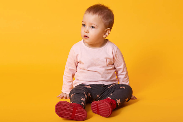 Little pretty girl sits on floor, dressed sweater, trousers with stars and red shoes, looks aside, finds friend for playing, wants on pens to mom. Copy space for your advertisment or promotion text - Foto, imagen