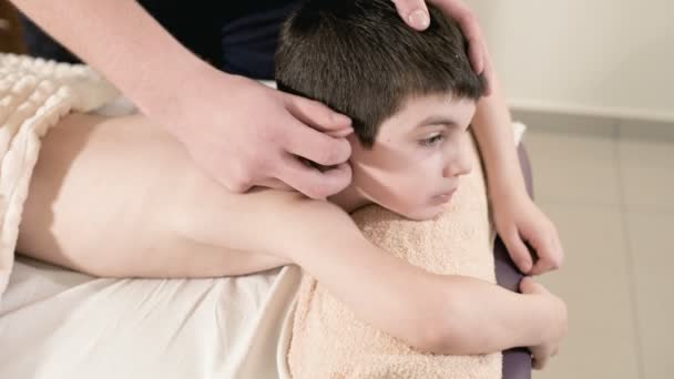 A male physiotherapist masseur makes a healing relaxing massage to a little smiling boy lying on a massage bed. Ear massage - Materiał filmowy, wideo